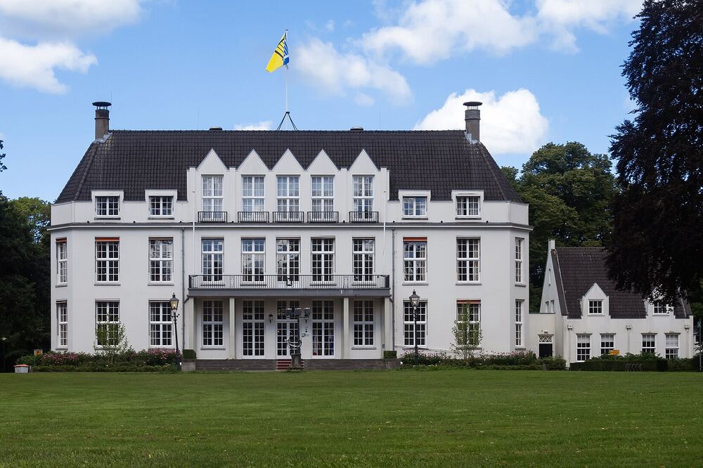 Het witte landhuis Jagtlust in Bilthoven, tegenwoordig het gemeentehuis van De Bilt, met puntgevels en balkons, een vlag op het dak en een breed gazon met oude bomen ervoor onder een blauwe lucht